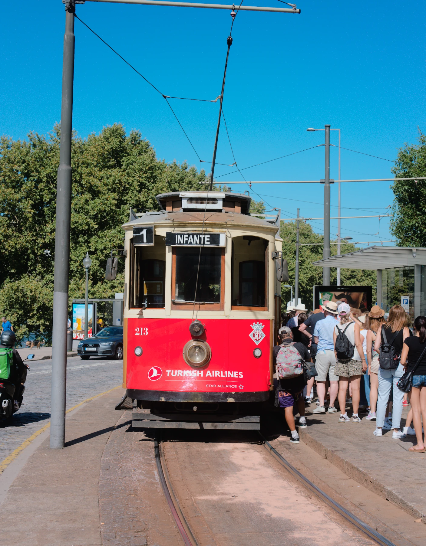 A red tram in Porto, with blue sky, green trees and people waiting to board, rendered with Everyday